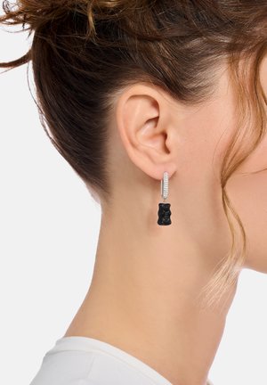 Schwarzes Gummibärchen-Ohranhänger mit einem funkelnden silbernen Reifen. Der Bär ist mit einem glänzenden Finish detailliert und hängt unter dem Ohr.