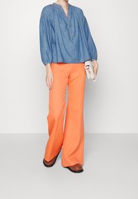 Blusa in denim azzurro con chiusura a bottoni e maniche arricciate, abbinata a pantaloni arancioni a vita alta e gamba larga, con calzature marroni.