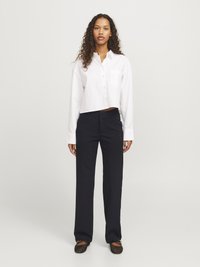 Femme debout, portant une chemise blanche à manches courtes boutonnée, un pantalon noir droit et des ballerines noires, sur un fond léger uni.