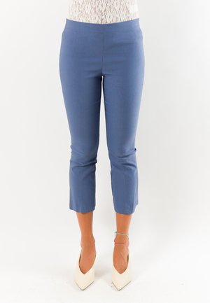 Donna che indossa pantaloni blu corti e tacchi bianchi a punta, in piedi davanti a uno sfondo bianco uniforme.