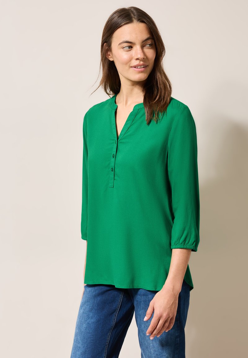 Camisa verde de manga larga con escote en V y botones en la parte frontal. Hecha de una tela suave, cuenta con puños fruncidos y un corte relajado.