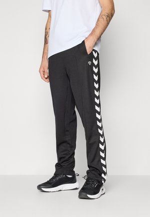 Svarta träningsbyxor med en slim fit, med vita chevronränder längs sidorna och fickor med dragkedja. Kombinationen är med svarta sneakers.