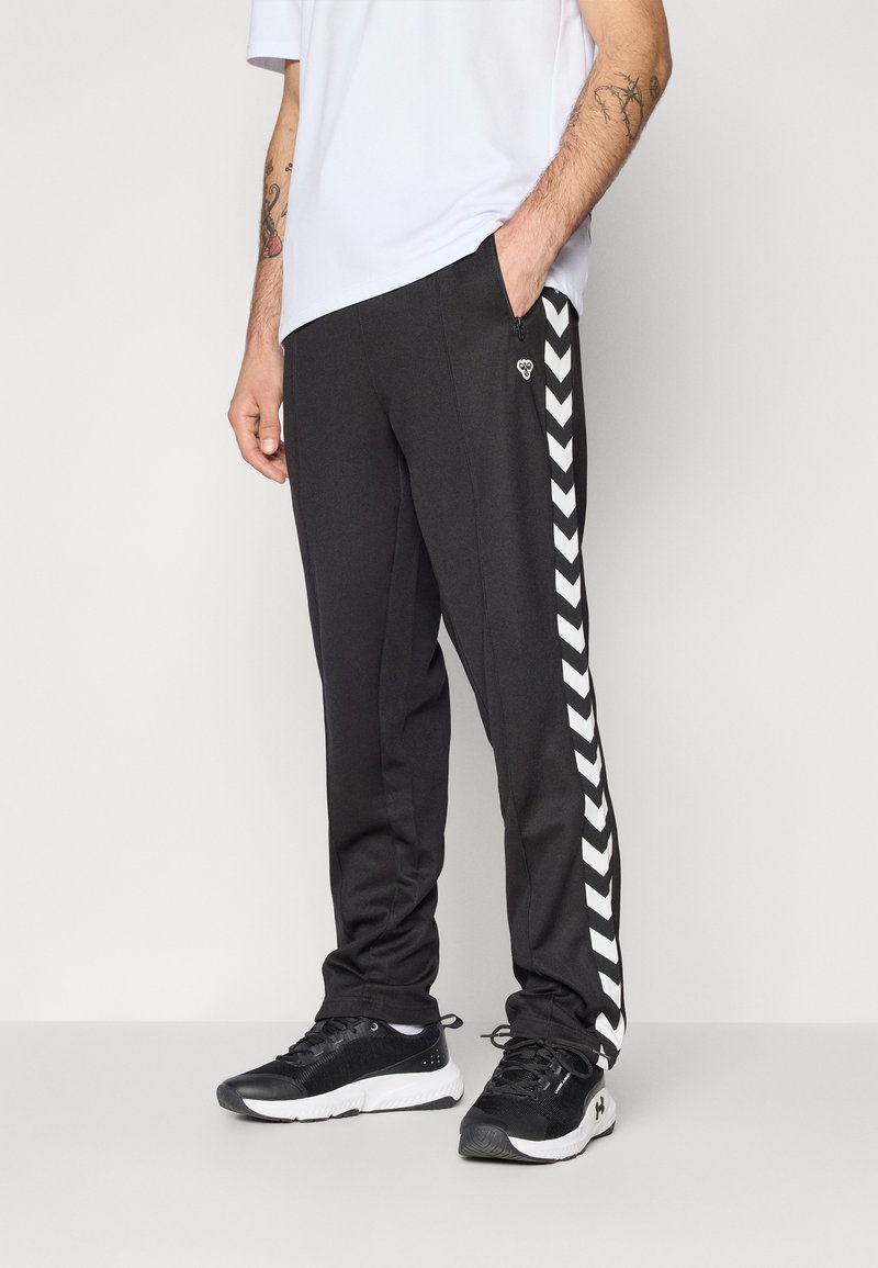 Čierne atletické nohavice so slim fit strihom, ktoré majú biele chevron pruhy po stranách a vrecká na zips. Kombinované s čiernymi teniskami.