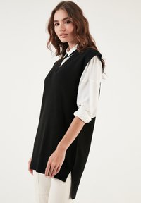 Black sleeveless knit top with a V-neck, side slits, and a relaxed fit. Worn over a white button-up shirt. Smooth texture.