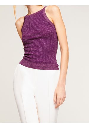Woman wearing a sleeveless, ribbed, purple metallic knit top paired with high-waisted, white tailored trousers against a plain background.