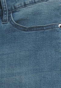 Denimová látka v světle modré barvě s jemnou strukturou, obsahující přední kapsu a detail stříbrného knoflíku na pase.