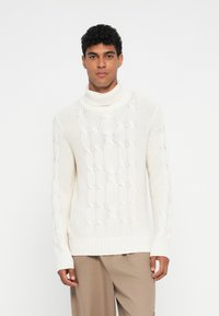 Suéter blanco de punto trenzado con un alto cuello acanalado. La textura es suave, con patrones en relieve, complementado por mangas largas y un corte relajado.