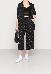 Schwarzer Blazer mit Punkten über einem schwarzen Cropped-Top und passenden Culottes, kombiniert mit weißen Sneakers und einer gewebten schwarzen Tasche.
