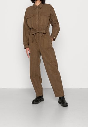 Woman wearing a brown buttoned jumpsuit with a waist tie, long sleeves, chest pockets, and black ankle boots, standing against a plain background.