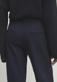 Tummasti neulottu villapaita, jossa on rento malli, yhdistettynä tummansinisiin räätälöityihin housuihin, joissa on kaksi takataskua ja sileä pinta.