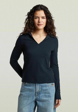 Femme aux cheveux bouclés foncés portant un haut noir à manches longues et col en V, et un jean bleu clair, debout devant un fond clair uni.