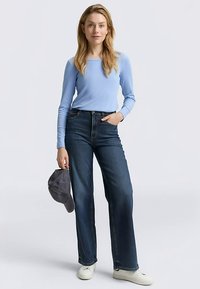 Ljusblå långärmad skjorta, mörka denimbraxor med raka ben, vita sneakers och grå keps hålls i vänster hand.