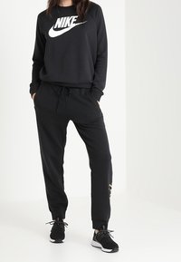 Svart bomullströja med en vit Nike-logga, tillsammans med svarta mjukisbyxor med sidofickor och dragsko, bärs med svarta sneakers.