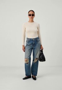 Beige long-sleeve top, denim jeans with large rips, black sunglasses, and black handbag. Flat black shoes complete the outfit.