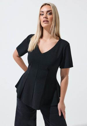 Blonde woman wearing a black short-sleeve V-neck top with flared hem and black pants, standing against a plain white background.