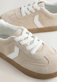 Beige suede sneakers with white laces and side accents. Features a rubber sole with a textured grip and stitched detailing.