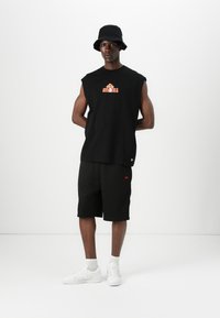 Black sleeveless shirt with orange graphic, paired with black mesh shorts featuring a red logo. Worn with white shoes and a black bucket hat.