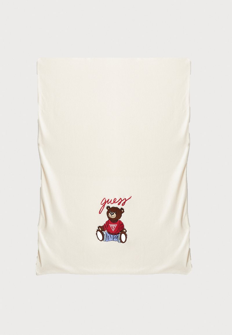 Couverture couleur crème ornée d'un ourson brodé portant un sweat à capuche rouge et un jean bleu, avec le mot "guess" en rouge en lettres cursives.
