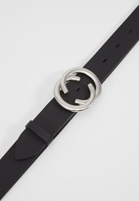Zwarte leren riem met een zilveren ronde gesp. De band heeft gaten voor aanpassing en een lus voor veilige bevestiging.