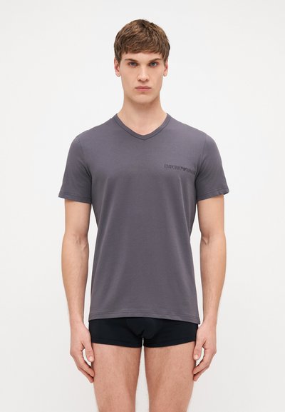 Šedé tričko s výstřihem do V z jemné látky, krátké rukávy, s malým černým logem na levé straně. Model má na sobě černé kraťasy.