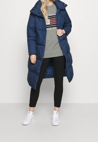Abrigo de plumas azul marino con un alto cuello, textura ligeramente brillante, que cuenta con un cierre de cremallera y bolsillos laterales, combinado con un sudadera gris y leggings negros.