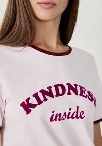 Donna che indossa una maglietta bianca con bordi bordeaux e le parole "KINDNESS inside" stampate in grandi e bold lettere bordeaux.