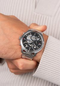 Rustfrit stål ur med sort urskive, sølvfarvede detaljer, chronograph-funktioner og en led-armbånd, båret på et håndled med lys hudtone.