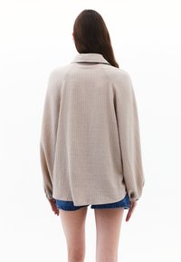 Giacca beige con un tessuto testurizzato, colletto e vestibilità ampia. Presenta maniche arricciate e orlo arrotondato, indossata sopra pantaloni di jeans corti.