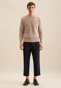 Beige geripptes Strickoberteil kombiniert mit dunkelblauen maßgeschneiderten Hosen und schwarzen Schnürschuhen. Einfache, klare Linien und strukturierter Stoff.