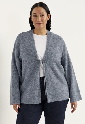 Vrouw met een zachte, grijze cardigan met lange mouwen, voorgebonden over een wit overhemd en donkere broek, staand tegen een witte achtergrond.