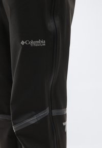 Μαύρο αδιάβροχο παντελόνι με λογότυπο Columbia Titanium και αντανακλαστικές ρίγες κατά μήκος μιας κατακόρυφης ραφής στο πόδι.