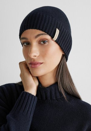 Stickad marinblå mössa med ribbad textur och en sidolapp. Bärs bekvämt, matchande med en polotröja, vilket framhäver värme och enkelhet.