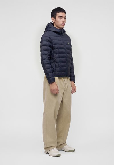 Jeune homme debout portant une doudoune noire à capuche, un pantalon large beige et des baskets claires sur un fond uni.