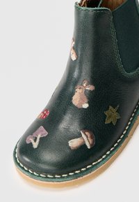 Botín de tobillo de cuero verde oscuro con detalles bordados de conejos, setas y hojas, con suela de goma natural y bordes cosidos.