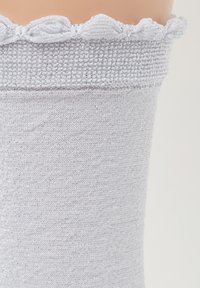 Světlá šedá ponožka vyrobená z texturované pleteniny. Horní část má volánkový okraj se stříbrným třpytem. Hladké tělo s jemným leskem.