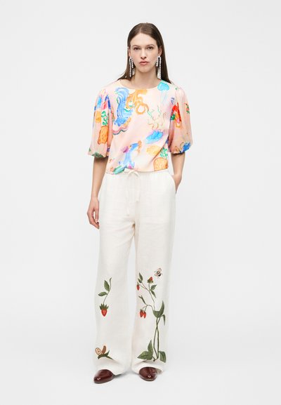 Femme portant un chemisier à manches ballon imprimé de créatures marines colorées et un pantalon blanc à jambes larges avec broderie botanique, accompagné de chaussures marron.