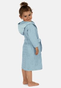 Helleblaue Kapuzenrobe aus Plüschmaterial, mit einem Gürtel, langen Ärmeln und strukturiertem Stoff. Kind steht barfuß da und lächelt leicht.