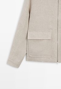 Beige textured jacket with long sleeve, front flap pocket, and silver zipper closure on the right side.