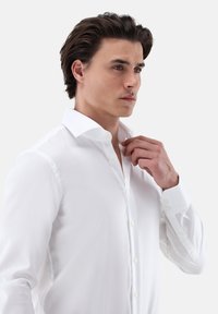 Weiße Hemdbluse mit weiter Kragenlinie, glatter Textur und langen Ärmeln. Verfügt über einen taillierten Schnitt und dezenten Glanz.