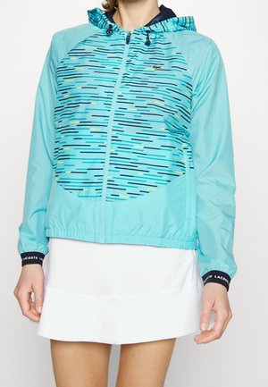 Vrouw draagt een lichtblauwe capuchonvest met donkerblauwe horizontale strepen en een witte rok, handen ontspannen langs het lichaam.