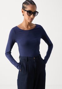 Langermet navy topp i glatt stoff, kombinert med mørke teksturerte høyt liv bukser. Modellen har på seg store svarte solbriller.
