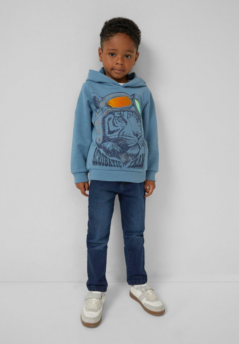 Jongentje in een blauwe hoodie met een tijger- en brilprint, donkere jeans en witte sneakers, staand tegen een grijze muur.