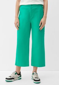 Grüne, weit geschnittene Hosen aus glattem Material mit Knopfverschluss und seitlichen Taschen, kombiniert mit schwarz-weißen Sneakers.
