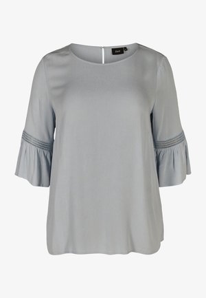 Blouse bleu clair avec un décolleté rond et des manches trois-quarts en forme de cloche. Présente des accents texturés au bord des manches et une fermeture à bouton au dos.