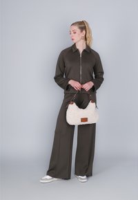 Braune Reißverschlussjacke, passende weit geschnittene Hose und flauschige weiße Handtasche mit braunen Akzenten. Das Model steht vor einem grauen Hintergrund.