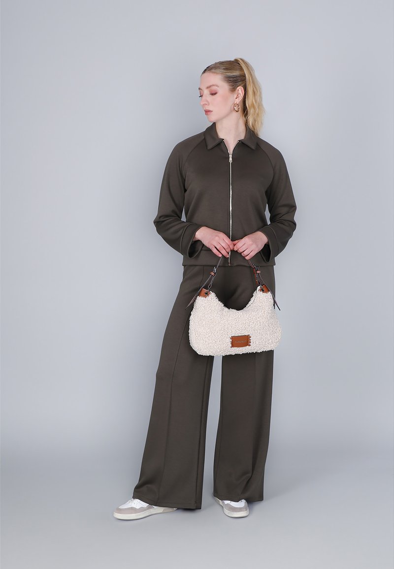 Braune Reißverschlussjacke, passende weit geschnittene Hose und flauschige weiße Handtasche mit braunen Akzenten. Das Model steht vor einem grauen Hintergrund.