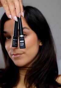 Deux tubes d'eyeliner noirs tenus dans une main, l'un étiqueté "Eyeliner Brillant" et l'autre "Eyeliner Coloré", tous deux avec des designs élégants.