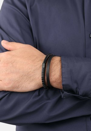 Black leather bracelet with smooth and textured bands, featuring round beads and a rectangular metal accent on a wrist.