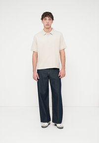 Μπεζ πόλο μπλούζα με κοντά μανίκια και γιακά, σε συνδυασμό με σκούρο μπλε τζιν καμπάνα. Το μοντέλο φοράει λευκά αθλητικά παπούτσια. Ουδέτερο φόντο.