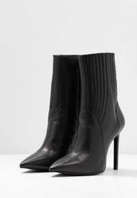 Pari mustat nahkaiset nilkkasukat teräväkärkisellä muotoilulla, jotka ovat korkeilla koroilla ja joissa on pystysuuntaista ompelua sivuilla valkoista taustaa vasten.